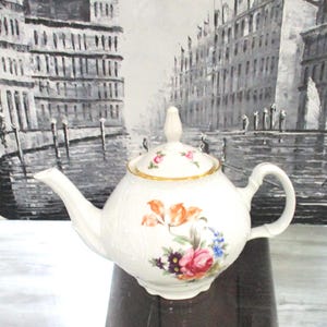 May include: White porcelain teapot with a floral design. The teapot features orange, pink, and blue flowers, a gold trim, and a matching lid with a small floral pattern. The handle and spout are curved.