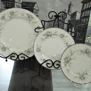 Pode incluir: Três pratos de porcelana branca com um design floral e acabamento prateado são exibidos em um suporte de metal preto. Os pratos são organizados em forma de cascata, com o prato maior na parte inferior e o prato menor na parte superior.