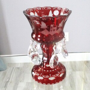 May include: A deep red glass vase featuring a scalloped rim and etched white patterns of trees and buildings. The vase has a pedestal base with circular motifs and is adorned with multiple clear, faceted crystal teardrop prisms hanging from the bowl.