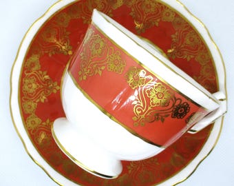 May include: A white teacup and saucer with a coral red and gold floral pattern. The saucer has a scalloped edge.