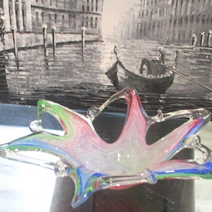 May include: A multi-coloured glass bowl in a starburst shape. The bowl has red, green, and blue swirls, with clear glass details. The background is a black and white painting of a canal scene with a gondola.