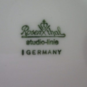 以下が含まれることがあります： 白い磁器の表面のクローズアップ。緑色の文字で「Rosenthal studio-linie I GERMANY」と書かれています。ローゼンタールのロゴはブランド名の上に王冠があります。テキストはセリフ体です。