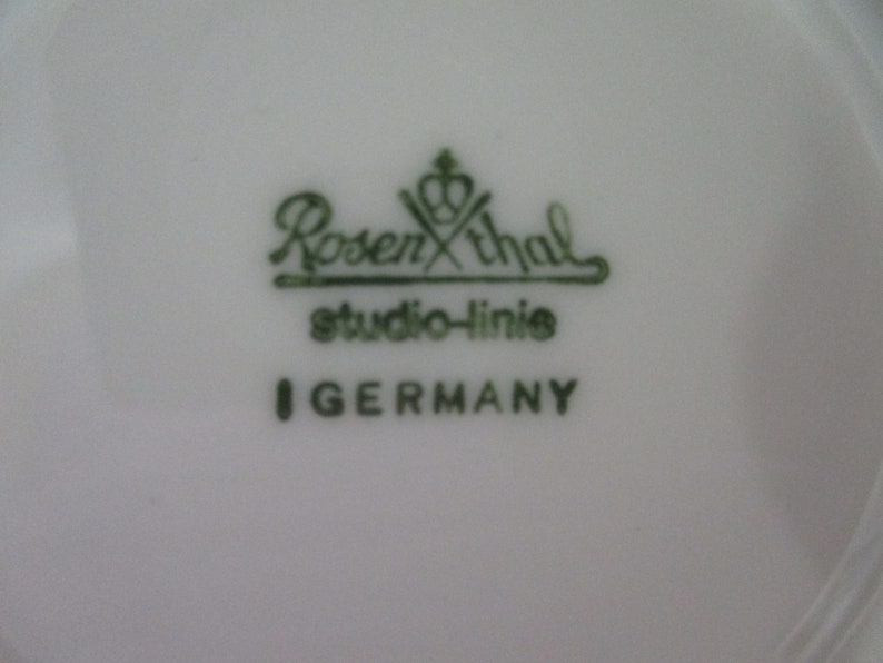 以下が含まれることがあります： 白い磁器の表面のクローズアップ。緑色の文字で「Rosenthal studio-linie I GERMANY」と書かれています。ローゼンタールのロゴはブランド名の上に王冠があります。テキストはセリフ体です。