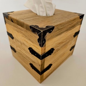 May include: Wooden tissue box with a square shape and a natural wood grain finish. The corners are reinforced with decorative black metal brackets. White tissues are visible at the top.