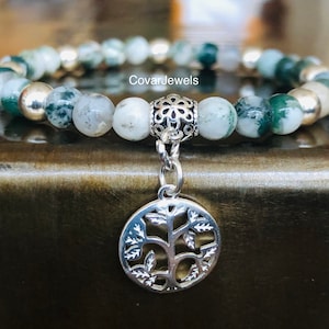 May include: A green and white agate stone bracelet with a silver tree of life charm. The bracelet has a silver clasp and is on a wooden surface. CovarJewels is printed on the bracelet.