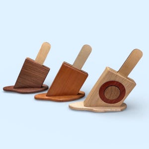 May include: Three wooden phone stands in the shape of melting ice lollies. The stands are made from various woods, including dark brown, medium brown, and light wood with a red circle. Each stand features a wooden stick.