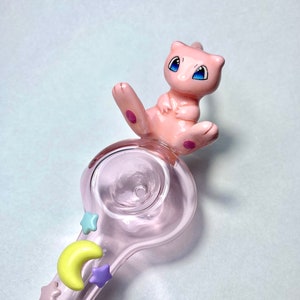 May include: A pink glass pipe with a cartoon Mew figure on top. The pipe features a clear bowl and a stem adorned with pastel-colored stars and a crescent moon. The text "Blessed DNA" is at the bottom.