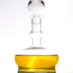 Könnte beinhalten: Eine klare Glas-Dekantierkaraffe mit rundem Verschluss, gefüllt mit einer goldenen Flüssigkeit. Die Karaffe hat einen bauchigen Boden mit horizontalen Rillen und einen langen Hals. Der Verschluss ist eine klare, runde Kugel.