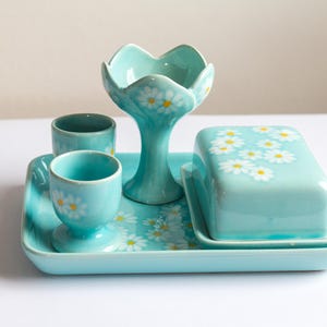 Può includere: Un set da colazione vintage in ceramica color acquamarina con vassoio, burriera, due portauova e un elemento di servizio con stelo. Il set è decorato con accenti di margherite bianche. Un'aggiunta affascinante a qualsiasi cucina.