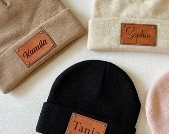 May include: Four beanies in tan, cream, black, and pink, each with a rectangular leather patch. The patches are embossed with the names "Kamila", "Sophie", "Tania", and "Kysten". The beanies are folded at the brim.