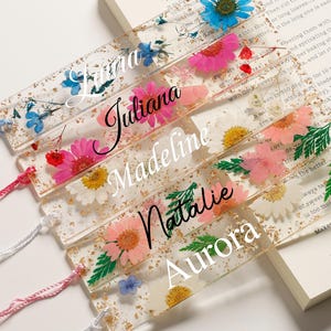 May include: Clear resin bookmarks with dried flowers and gold flakes. Each bookmark features a name in elegant script and a colorful tassel. The bookmarks are resting on an open book.