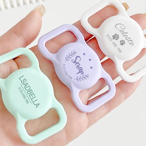 Può includere: Tre targhette identificative per animali domestici colorate: verde menta, lavanda e bianco. Ogni targhetta ha una forma arrotondata con un'apertura rettangolare. Le targhette sono personalizzate con nomi, numeri di telefono e impronte di zampe.