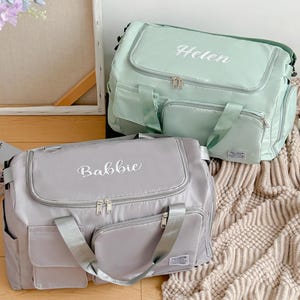 Peut inclure: Deux sacs de sport personnalisés. L'un est gris clair avec le nom "Babbie" brodé en blanc. L'autre est vert menthe avec "Helen" brodé en blanc. Les deux sacs ont plusieurs poches et des poignées.