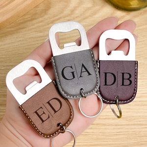 May include: Three personalised bottle openers with initials. The openers have a metal top and a leatherette bottom in brown, grey, and burgundy. Each has a keyring attached. The initials "ED", "GA", and "DB" are engraved on the leatherette.