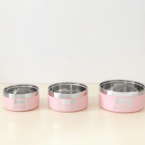 May include: Three pink pet bowls with stainless steel interiors. Each bowl has a silver bone design and the name "NANA" printed on the front. The bowls are in different sizes, suitable for various pet needs.