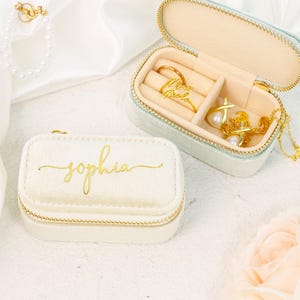 May include: An off-white jewellery box with the name "sophia" in gold script. The box is zippered and has a gold trim. An open, light blue jewellery box contains gold rings and a necklace. A pearl bracelet is also visible.