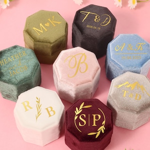 Pode incluir: Uma coleção de caixas de anel de veludo em várias cores, incluindo verde, rosa e cinza. Cada caixa octogonal apresenta letras douradas com iniciais ou datas. As caixas estão dispostas sobre uma superfície rosa, com detalhes florais.