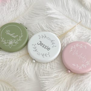 Può includere: Tre specchietti compatti personalizzati in colori pastello: verde, bianco e rosa. Ogni specchietto ha un nome in corsivo e un design floreale. Gli specchietti rotondi sono su un letto di piume bianche.