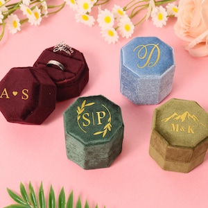 Pode incluir: Uma variedade de caixas de anel de veludo em várias cores, incluindo bordô, azul e verde. Cada caixa octogonal apresenta letras ou desenhos dourados, como iniciais, uma letra ou uma cordilheira. As caixas são exibidas em uma superfície rosa com detalhes florais.