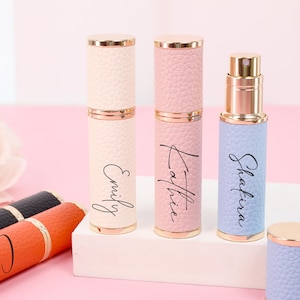 May include: A selection of pastel-coloured perfume atomisers. The cylindrical atomisers have gold accents and feature names such as "Emily", "Kathie", and "Shafira" in script. They are displayed on a white surface against a pink backdrop.