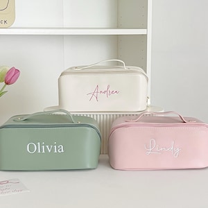 May include: Three personalized cosmetic bags in pastel colors: green, pink, and off-white. Each bag has a handle and a zipper closure. The names "Olivia", "Lindy", and "Andrea" are embroidered on the front.