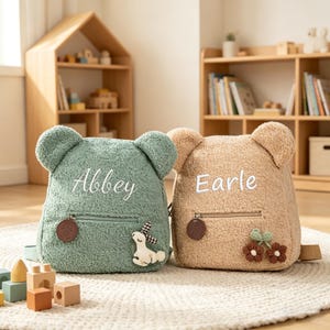 May include: Two plush bear-shaped backpacks, one green and one tan. The green backpack has the name "Abbey" embroidered on it, and the tan backpack has the name "Earle". Both have bear ears, a front pocket, and a decorative charm.