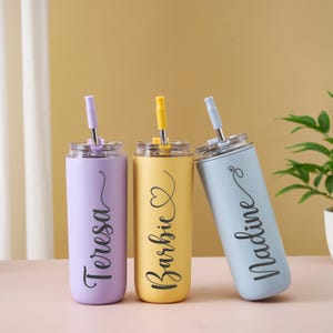 May include: Three personalized stainless steel tumblers in pastel colors: lavender, yellow, and light blue. Each tumbler has a matching straw and lid, with a name in a cursive font. The names are Teresa, Barbie, and Nadine.