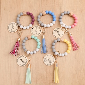 May include: A selection of keyrings with beaded bracelets in various colours, including grey, purple, blue, pink, mint green, and yellow. Each keyring features a wooden tag with a name and a tassel. The names on the tags are Chloe, Maria, Sarah, Vesta, and Daisy.