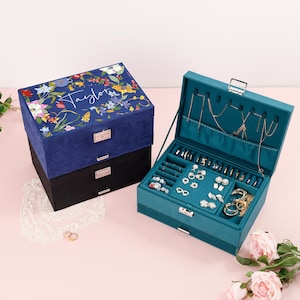 May include: A blue and black jewelry box set. The top box is navy blue with a floral design and the name "Taylor" in script. The open teal box displays jewelry, including necklaces, rings, and earrings. The boxes have silver-tone hardware.
