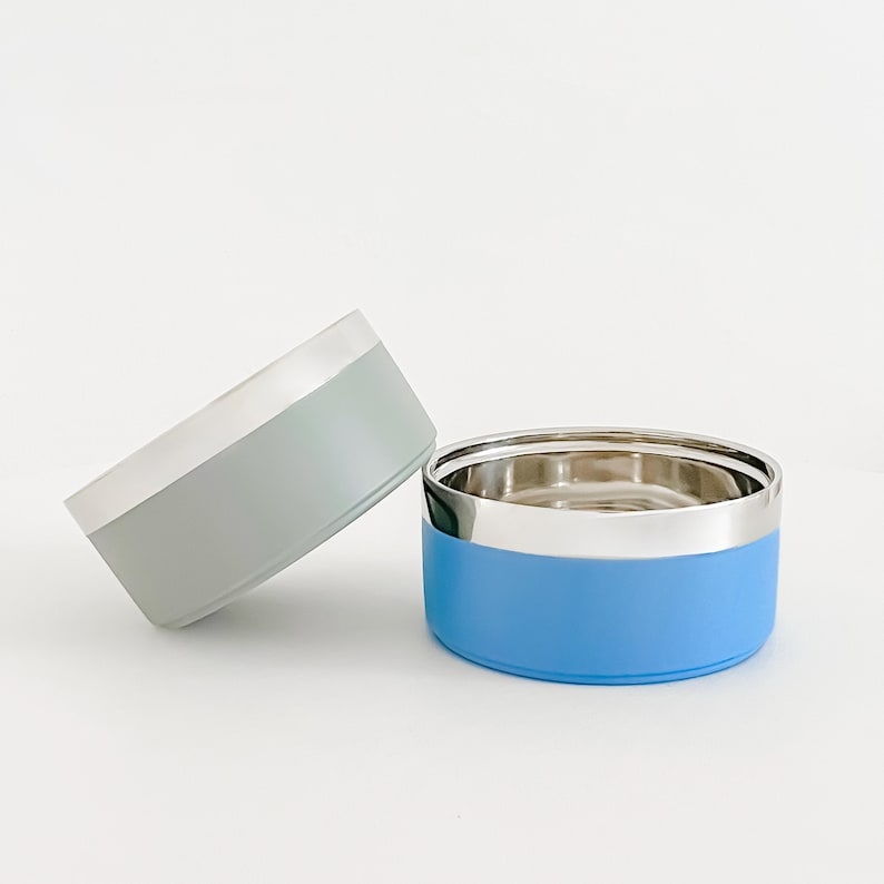 May include: Two pet bowls, one light blue and one gray, with a silver rim. The light blue bowl is upright, while the gray bowl is tilted. Both bowls have a stainless steel interior.