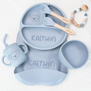 May include: A baby feeding set in light blue, featuring a divided plate, bowl, bib, cup, fork, and spoon. The set includes a koala bear graphic and the name "KAITHYN" printed on the plate and bib. The spoon and fork have wooden handles.