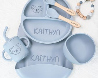 May include: A baby feeding set in light blue, featuring a divided plate, bowl, bib, cup, fork, and spoon. The set includes a koala bear graphic and the name "KAITHYN" printed on the plate and bib. The spoon and fork have wooden handles.