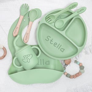 May include: A light green baby feeding set featuring the name "Stella" on the plate, bib, and utensils. The set includes a divided plate, a bib, a cup, a fork, and a spoon. The utensils have wooden handles. A pacifier clip is also included.
