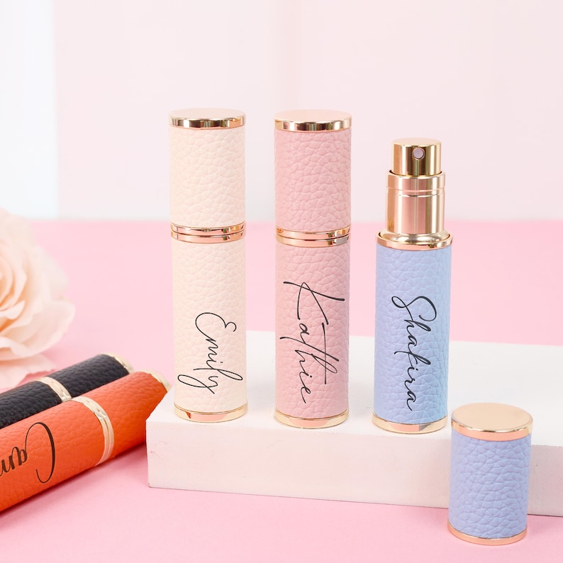 May include: A collection of personalized perfume atomizers in pastel colors. The atomizers are cylindrical with gold accents and feature names like "Emily," "Kathie," and "Shafira" in script. The atomizers are displayed on a white surface against a pink backdrop.