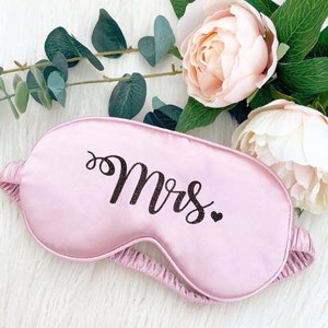 Puede incluir: Una m&aacute;scara de dormir de sat&eacute;n rosa con la palabra "Mrs." en letras de purpurina negra.