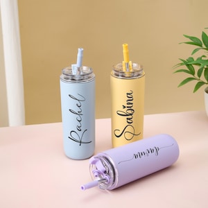 May include: Three personalized stainless steel tumblers in pastel colors: light blue, yellow, and lavender. Each tumbler has a matching straw and the name "Rachel", "Sabina", and "Jasmina" in a cursive font. The tumblers are on a light pink surface.
