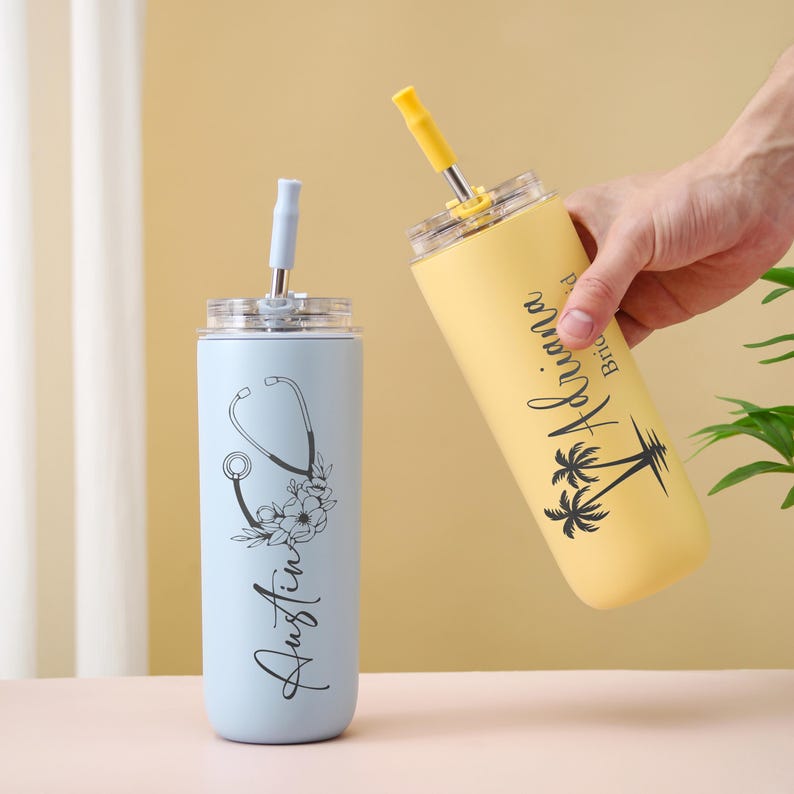 May include: Two stainless steel tumblers with straws. One is light blue with a stethoscope and floral design, the name "Austin" is written vertically. The other is yellow with the name "Ariana" and palm trees.