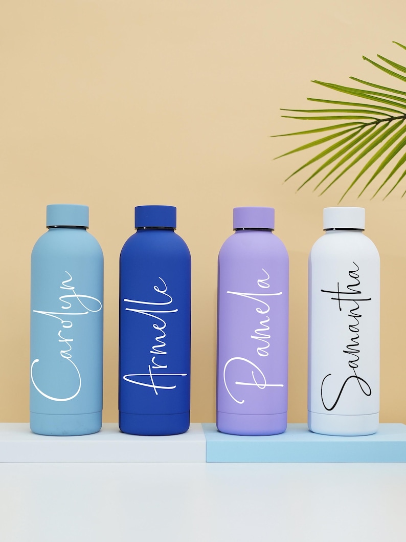 May include: Four personalized water bottles in blue, purple, white, and blue. The bottles are lined up on a light blue surface. The names "Carolyn", "Armelle", "Pamela", and "Samantha" are written in white on the bottles.