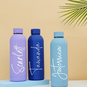 May include: Three personalized water bottles with names "Scarlett", "Tawanda", and "Taneerica" written in white script on a light purple, dark blue, and light blue bottle, respectively. The bottles are sitting on a light blue surface.