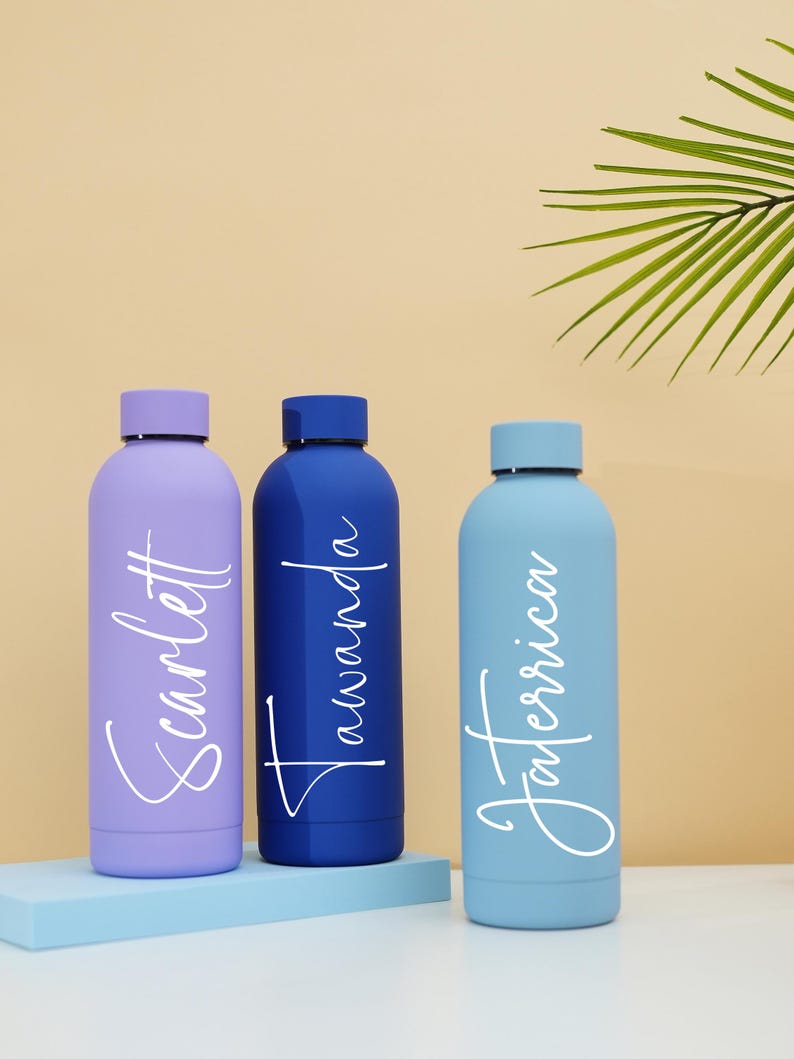May include: Three personalized water bottles with names "Scarlett", "Tawanda", and "Taneerica" written in white script on a light purple, dark blue, and light blue bottle, respectively. The bottles are sitting on a light blue surface.