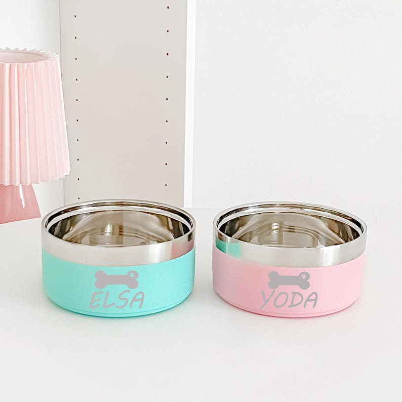 May include: Two stainless steel pet bowls with a light blue and pink base. The light blue bowl has the name "ELSA" and a bone graphic, while the pink bowl has "YODA" and a bone graphic. Both bowls have a silver rim.