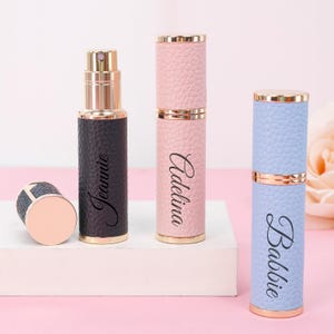 May include: A collection of three refillable perfume atomizers in black, pink, and blue, with gold-colored accents. The black atomizer is labeled "Jeannie", the pink one "Adelina", and the blue one "Babbie". One atomizer is disassembled, showing the inner bottle.
