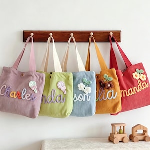 Peut inclure: Plusieurs sacs fourre-tout en velours côtelé colorés, chacun brodé d'un nom. Les sacs, dans des tons de violet, vert, bleu, orange et rouge, sont décorés d'ornements uniques et sont suspendus à un porte-manteau en bois.