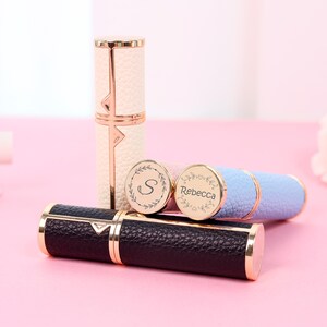 May include: A collection of personalized perfume atomizers in various colors, including black, white, and blue. Each atomizer features gold-colored accents and a textured leather-like finish. Some have engraved initials or names.
