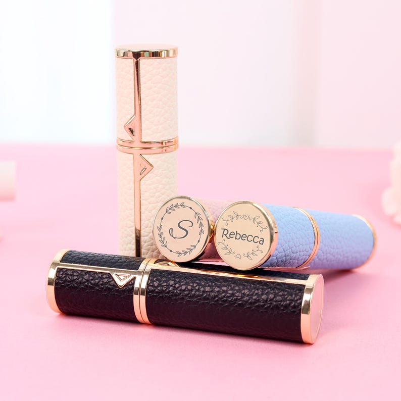 May include: A collection of personalized perfume atomizers in various colors, including black, white, and blue. Each atomizer features gold-colored accents and a textured leather-like finish. Some have engraved initials or names.
