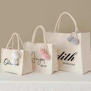 May include: Three off-white canvas tote bags with white handles. Each bag has a different name in black script. The bags are decorated with fluffy pom-poms in white, pink, and gray. The middle bag says "Anger Bride". The largest bag says "dith Bridesmaid".
