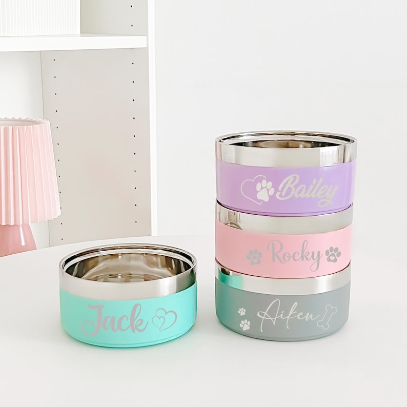 May include: Stainless steel pet bowls in pastel colors: mint green, pink, lavender, and gray. Each bowl has a silver rim and is personalized with a pet's name and paw print design. The bowls are stacked and arranged on a white surface.