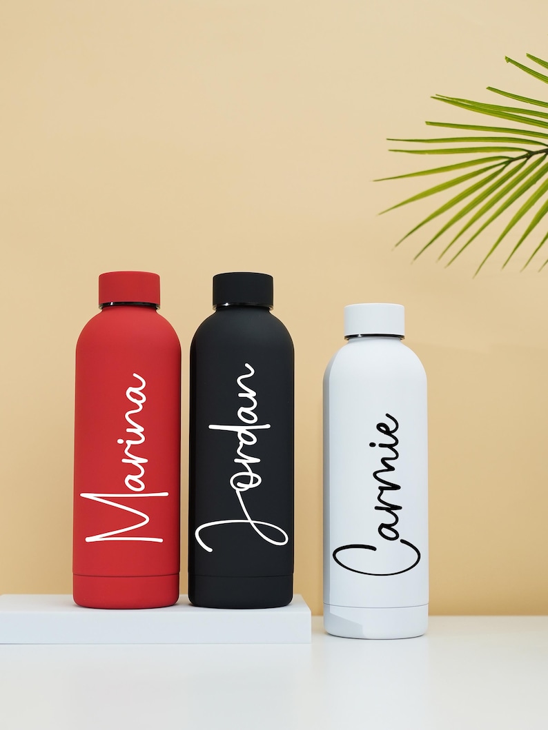 May include: Three stainless steel water bottles with personalized names: Marina, Jordan, and Cammie. The bottles are red, black, and white.