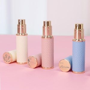 Puede incluir: Tres atomizadores de perfume personalizados en crema, rosa y azul con detalles dorados. Cada atomizador tiene una funda de polipiel texturizada y una tapa a juego con texto personalizado. Las tapas están sobre una superficie rosa.