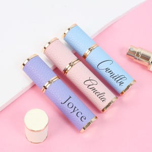 May include: Three personalized perfume atomizers in pastel colors: lavender, pink, and blue. Each atomizer has a gold-colored band and is inscribed with a name: Joyce, Amelia, and Camilla. A detached cap sits nearby.